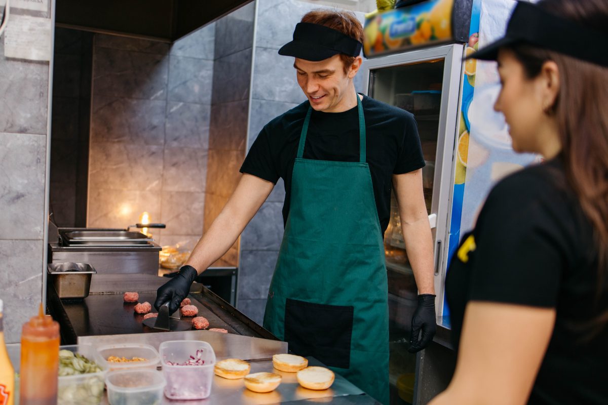 A young cook in a green apron working burgers on a flat-top grill while a coworker stands beside him.