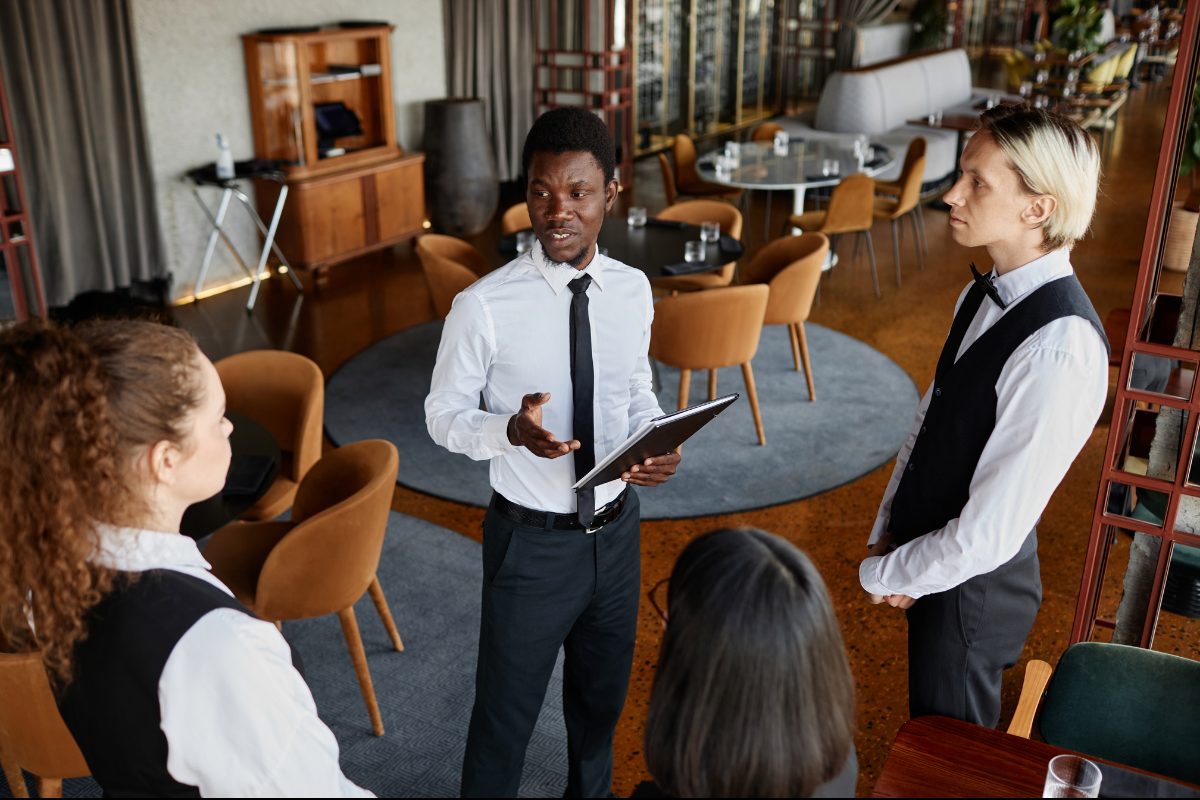 A restaurant manager holding a folder, leading a pre-shift huddle with servers in vests on the dining room floor.