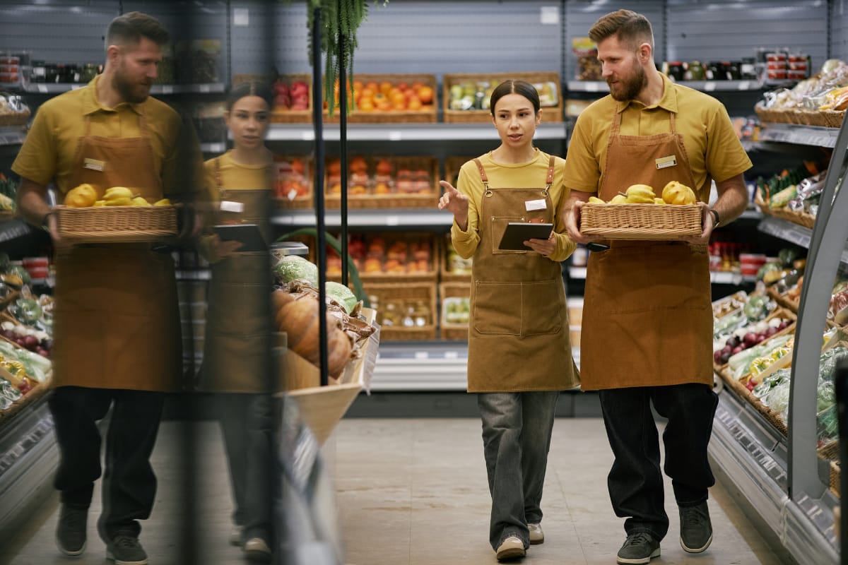 Two grocery associates in aprons walking a produce aisle together, one gesturing while holding a tablet, the other carrying a basket of fruit.