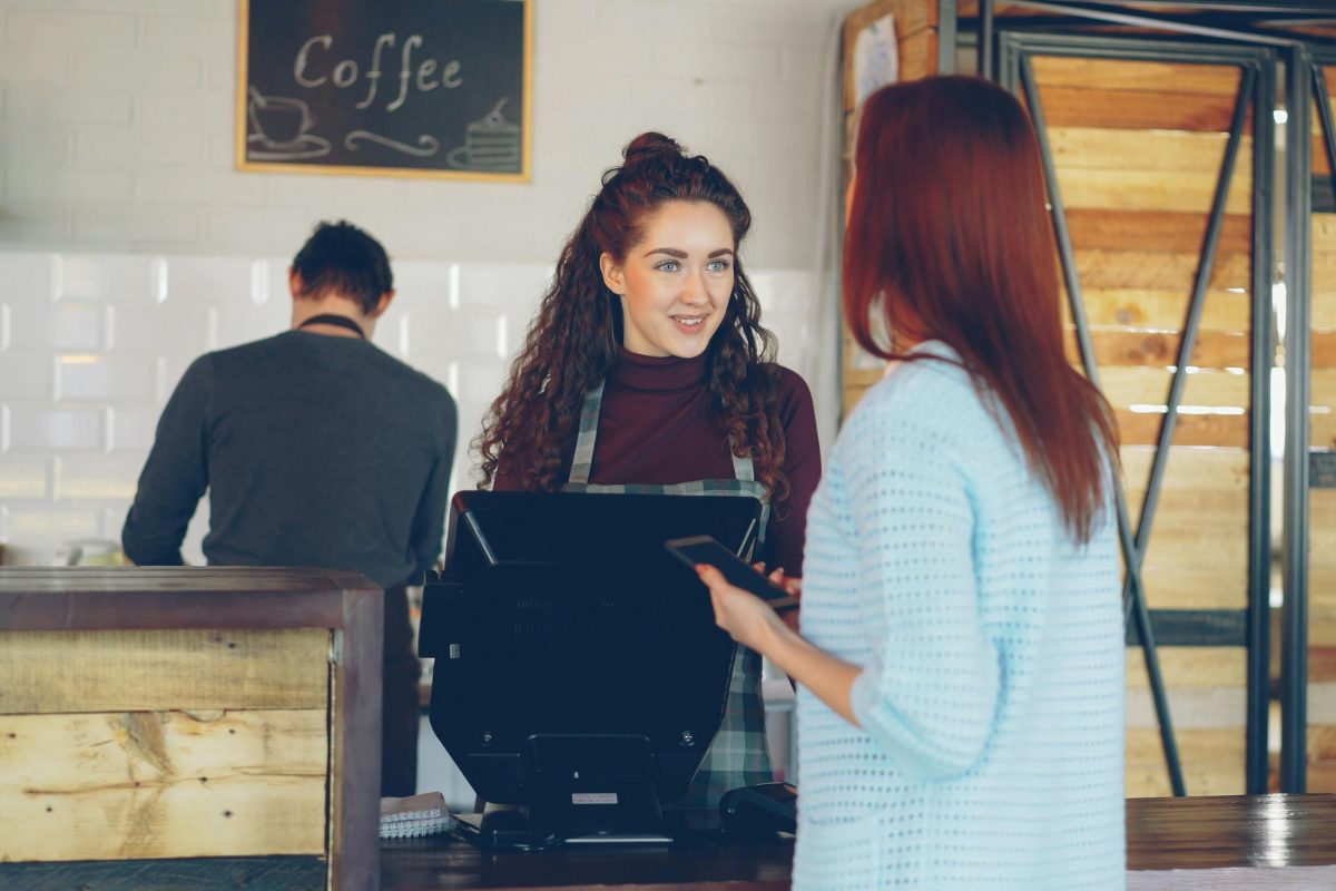 A counter worker in an apron smiling and talking with a customer at the register of a small coffee shop.