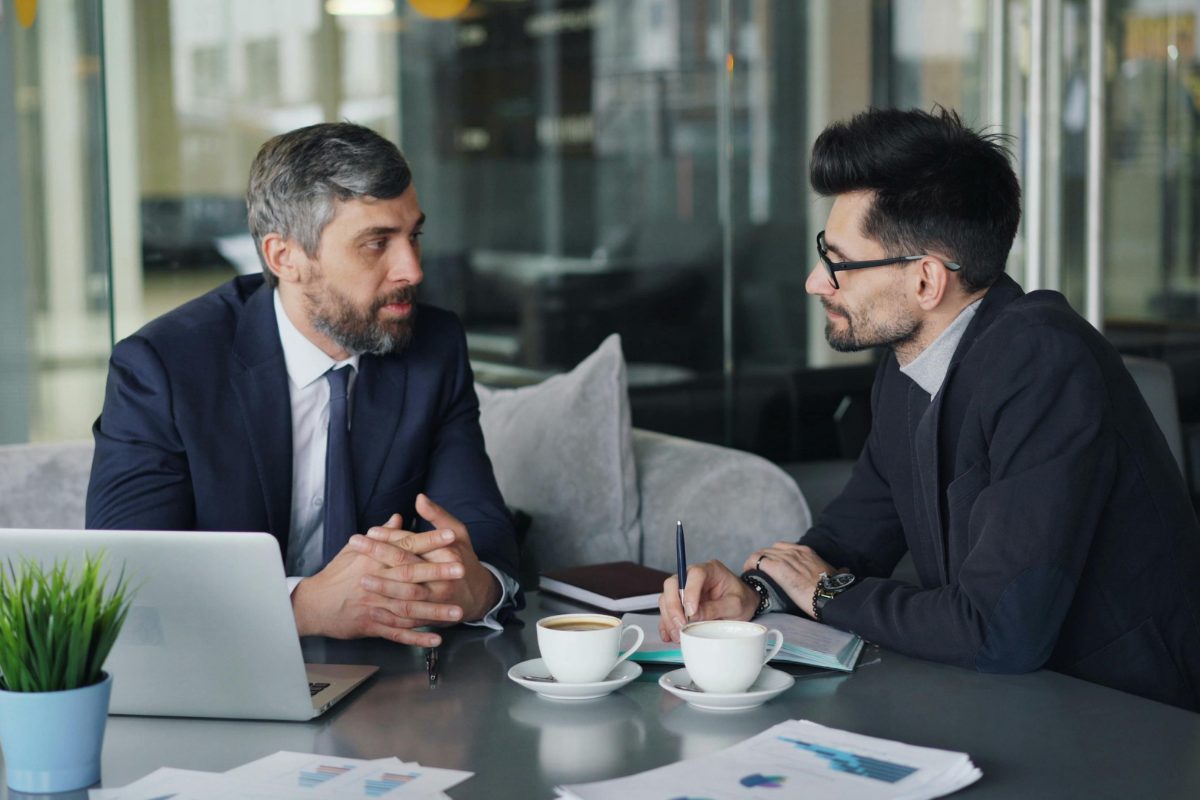 Two men in jackets at a table with coffee, a laptop and printed reports, one talking while the other takes notes.
