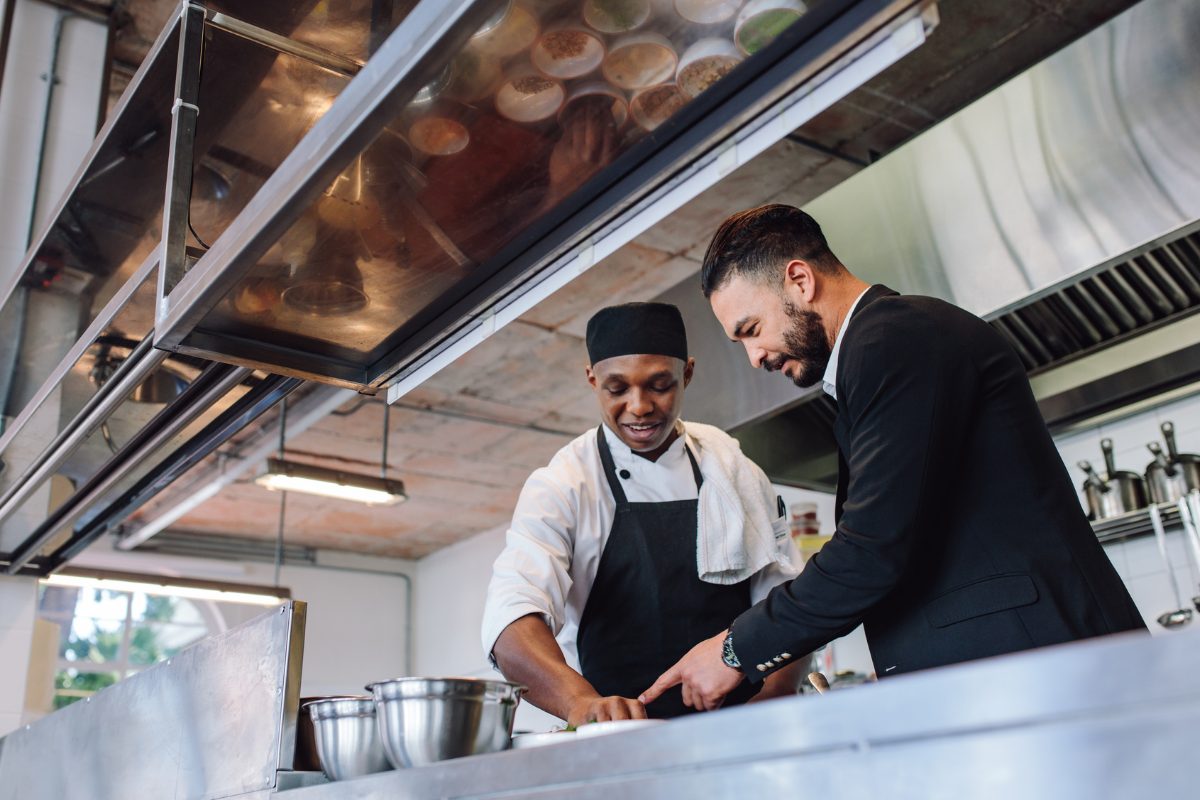 A manager in a blazer leaning over the pass with a cook in whites, both looking at a plate together.