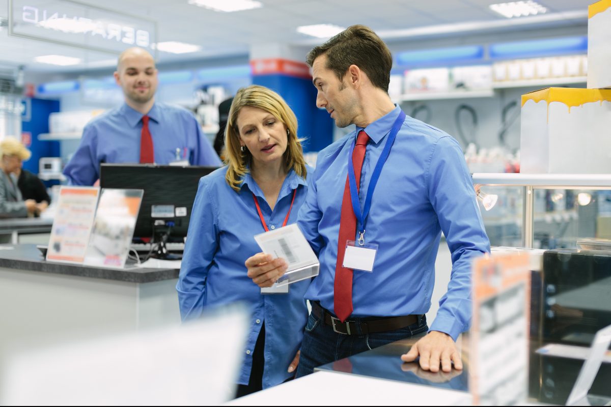Two electronics store employees in blue shirts and lanyards looking at a product tag together, a coworker behind them.