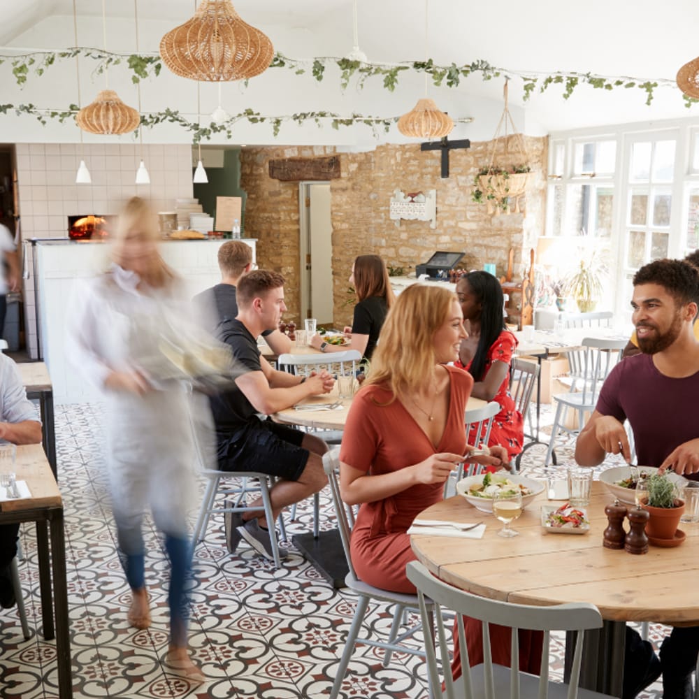 A busy sit-down restaurant floor at lunch, diners eating at wooden tables while a server crosses the room.