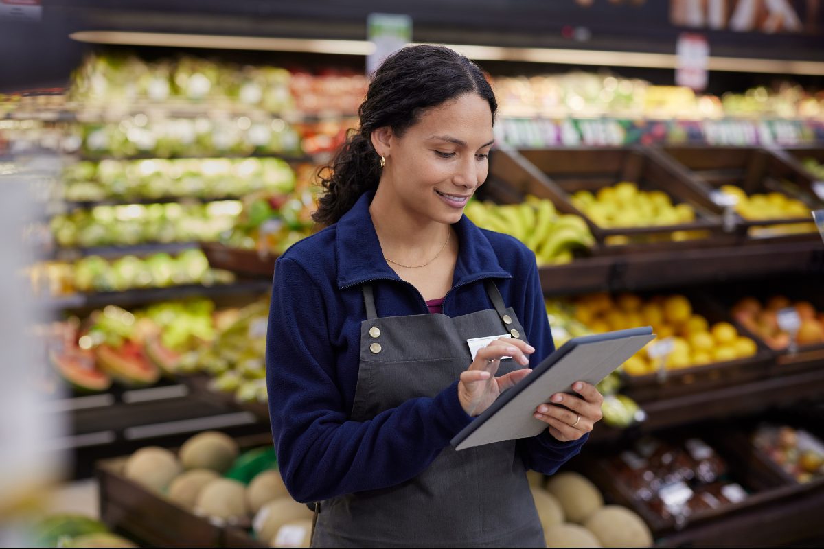 A grocery associate in an apron checking a tablet in the produce department, with fruit displays behind her.
