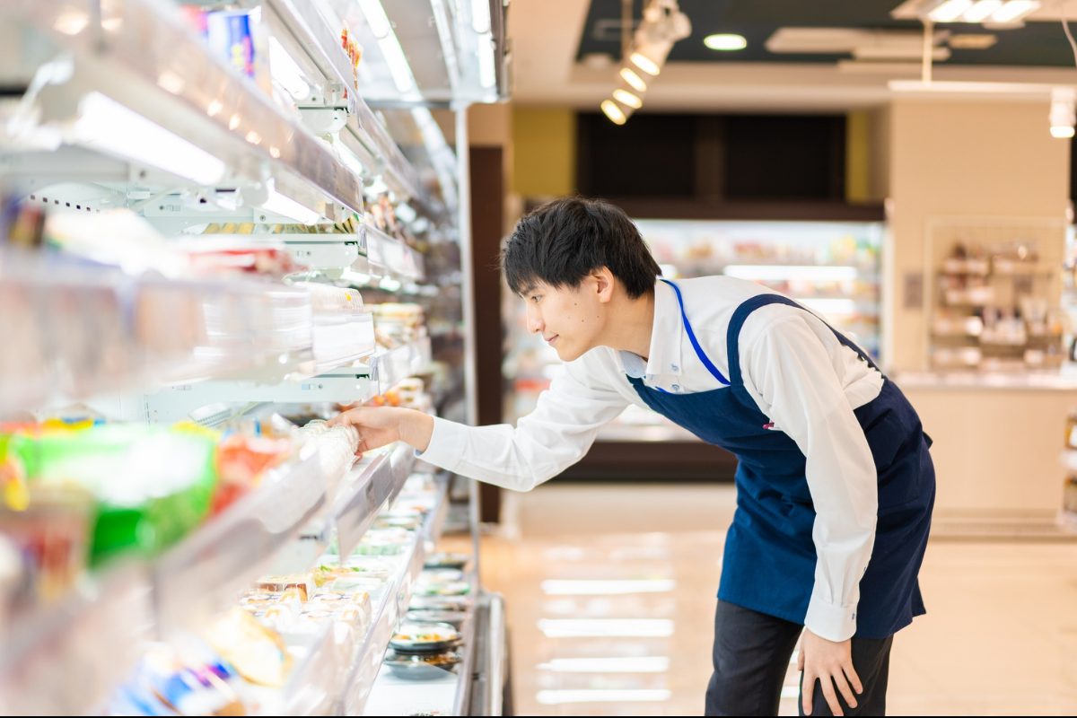 A convenience store associate in a blue apron leaning in to check a refrigerated grab-and-go case.