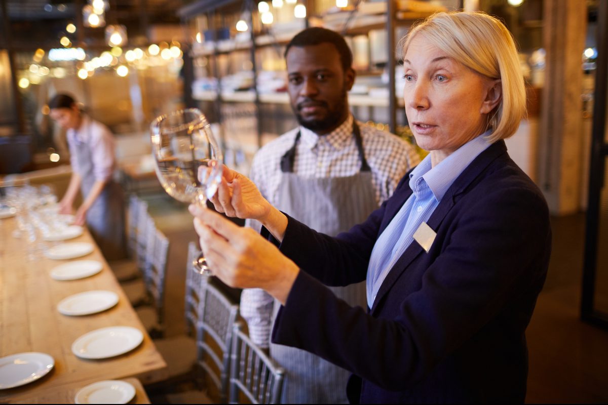A manager holding a wine glass up to the light while a server in an apron watches, a set table beside them.